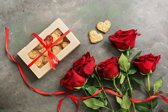 Cookies in a box in the shape of a heart, red roses and a red ribbon on a dark painted rustic background. Valentine's Day. Top view, flat lay.
