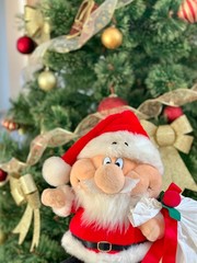 Soft, Christmas toy Santa on the background of the Christmas tree. Bright doll of Santa Claus with gifts with a bag. Preparation for the holidays, New Year's gifts for children.