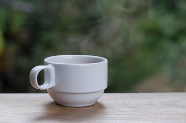 White hot coffee cup with smoke floating out Put on a wooden balcony With a blurred tree backdrop