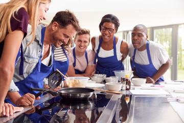 Male Teacher Making Pancake On Cooker In Cookery Class As Adult Students Look On