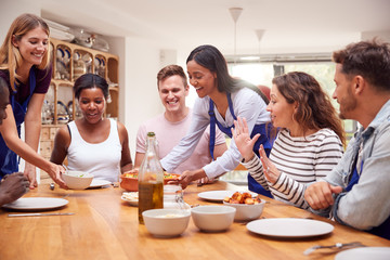 Group Of Men And Women Sitting Around Table Eating Meal They Have Prepared In Kitchen Cookery Class