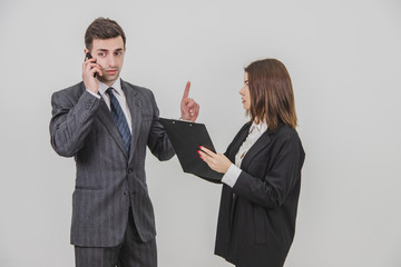Handsome busy businessman is talking on the phone, pointing his finger up for his secretary to wait. Secretery standing with clipboard, waiting for boss's signature.