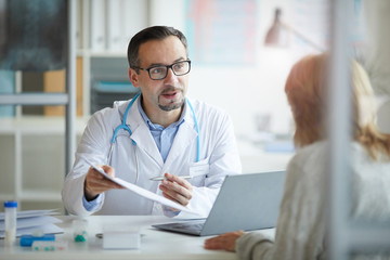 Mature male doctor in white coat pointing at document with treatment and showing it to his patient while they sitting at the table