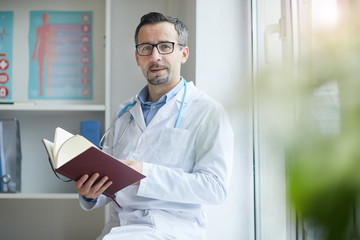 Portrait of mature male doctor in white coat sitting with note pad and checking his notes at office