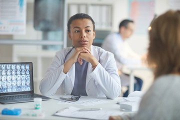 Serious female doctor sitting at her workplace and listening to complains of her patient at the...
