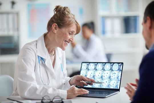 Mature female doctor sitting at the table and pointing at laptop with x-ray images and talking to the patient at office
