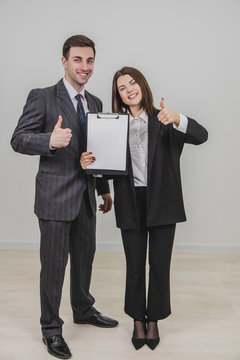 Successful business couple standing, holding together a clipboard with empty clear sheet of paper for text, giving thumbs up, smiling.