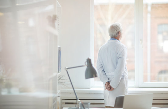 Rear View Of Senior Male Doctor In White Coat Standing Near The Window At Office Thinking And Resting After Work