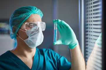 Fototapeta na wymiar Male doctor in medical cap eyeglasses and protective mask holding test tube in his hand and examining it