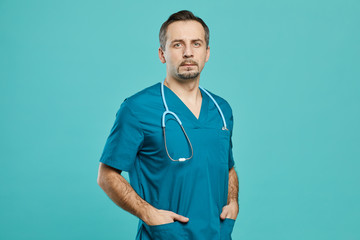 Portrait of mature male doctor in scrubs and stethoscope standing with hands in pocket and looking at camera against the blue background