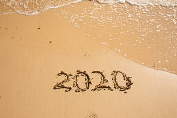 Summer beach holiday 2020 season golden sand - year - decade - new season lettering on the beach with wave and clear blue sea. Numbers 2020 year on the sea shore, message handwritten New Years concept