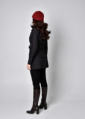 full length portrait of a pretty brunette girl wearing a black coat, leather boots and red beanie. Standing pose with back to the camera on a grey studio background.