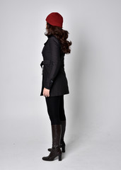 full length portrait of a pretty brunette girl wearing a black coat, leather boots and red beanie. Standing pose with back to the camera on a grey studio background.