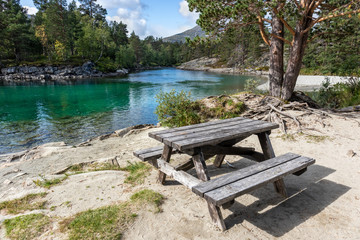 Fototapeta na wymiar Camping rest area for picnic with wooden table bench on sand by the cold sparkling river in pine forest mountains of Norway