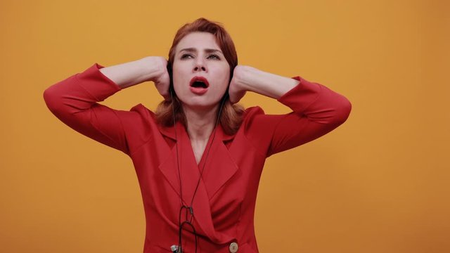 Caucasian scared young woman wearing fashion red jacket over isolated orange background having nice headphones, keeping hand on it, listening music. People lifestyle concept.