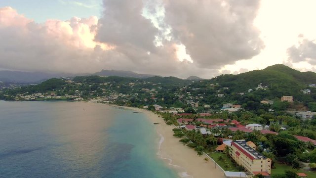 Slow aerial view of the coast of Grenada island, showing residential constructions, calm ocean waves on the sand seashore during dusk, beautiful clouds and lights