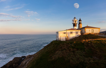 The double lighthouse of Punta Atalaia on the north coast of Spain in Galicia on the Atlantic. It is evening and sunset. Waves hit the coast and the moon is in the sky.