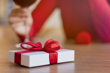 White gift and a ring in the box laying on the table. Young woman wearing a red sweater sitting with a glass of wine. Present with a red ribbon. Valentine's day and winter holidays gifts concept.