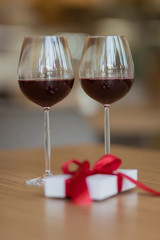 Two glasses with wine standing on a brown table at a restaurant. A white present giftbox with a red ribbon laying on the cafe's table. Valentine's day and winter holiday's concept.