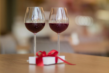 Two glasses with wine standing on a brown table at a restaurant. A white present giftbox with a red ribbon laying on the cafe's table. Valentine's day and winter holiday's concept.