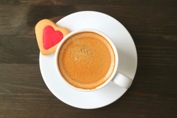 Top view of a cup of coffee with a heart shaped cookie on dark brown wooden table