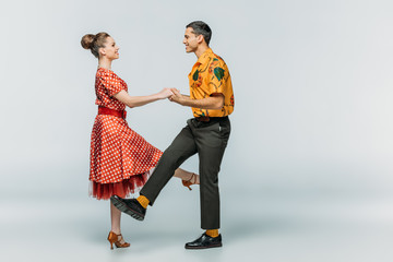 side view of smiling couple holding hands while dancing boogie-woogie on grey background