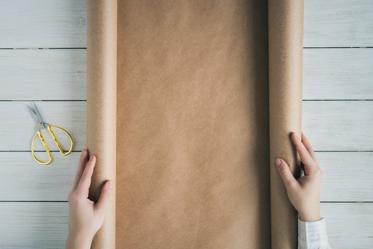 Female Hands Unfolding A Roll Of Wrapping Brown Craft Paper For Packing Gifts And Flowers On The Wooden White Table. Copy Space.