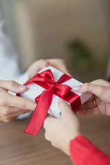 A small present is being passed by a couple from one to another. White little giftbox with a red ribbon in the hands. Valentine's day and winter holidays concept.