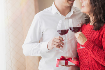 Young couple smiling to each other, drinking wine and holding white present with red ribbon in their hands at restaurant. Crossed wineglasses at a cafe, laugh and pass a gift. Valentine's day concept.