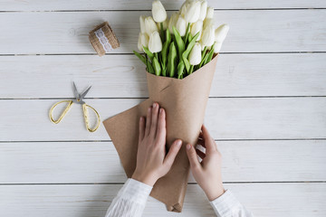 Woman florist wrapping beautiful bouquet of white tulips in pack craft paper on the wooden table....