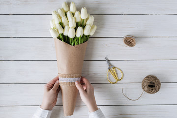 Woman florist wrapping beautiful bouquet of white tulips in pack craft paper on the wooden table. Flat lay