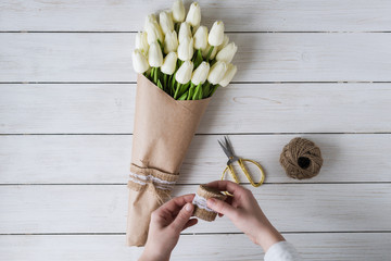 Woman florist wrapping beautiful bouquet of white tulips in pack craft paper on the wooden table....