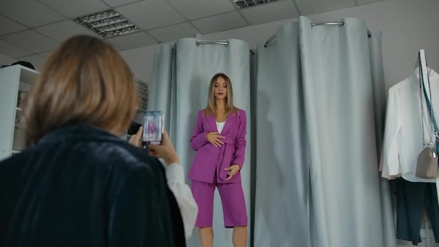 Low angle of young blonde female trying on trendy pink suit and spinning while friend taking picture with smartphone in clothing store, in slow motion.