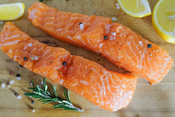 Two salmon or trout steaks lie on a wooden cutting board with fresh rosemary, chopped lemon, peppercorns and sea salt crystals. Horizontal orientation. Close-up.