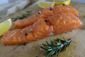 Two salmon or trout steaks lie on a wooden cutting board with fresh rosemary, chopped lemon, peppercorns and sea salt crystals. Horizontal orientation. Close-up.