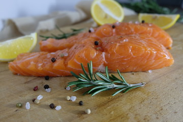 Two salmon or trout steaks lie on a wooden cutting board with fresh rosemary, chopped lemon, peppercorns and sea salt crystals. Horizontal orientation. Close-up.