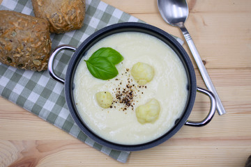 Cauliflower soup in a pot with checkered napkin and bun on wood. View from above