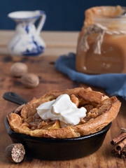 Homemade apple pie with caramel sauce and cream on a wooden table. Close-up, selective focus on the cream. Near a jar of caramel, milk and spices. Tasty dessert.