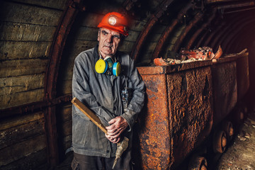An elderly man dressed in work overalls and a hard hat is standing near the old caravan. Mine worker. A tired miner in a coal mine looks at the light. Work in a coal mine. Portrait of a miner.
