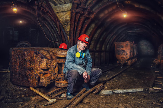 An elderly man dressed in work overalls and a hard hat is standing near the old caravan. Mine worker. A tired miner in a coal mine looks at the light. Work in a coal mine. Portrait of a miner.