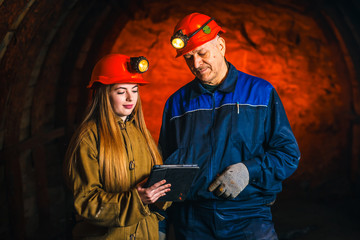 A beautiful young girl in a red helmet and with a electronic tablet in her hands is standing with a miner in a coal mine. Business plan discussion.