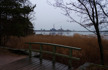 Wooden bridge on a walking path near the shore of a forest park on the island of Seurasaari and a view of the industrial city of Helsinki in Finland on a cloudy autumn evening