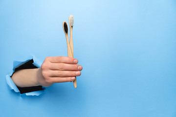 Female hand holding two bamboo eco toothbrushes through a torn blue paper wall. Dental care and Eco friendly and reuse concept. Copy space aside for your advertising content