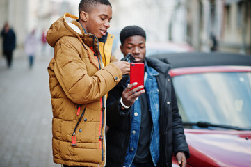 Two african male friends standing near red american convertible car and making selfie on phone.