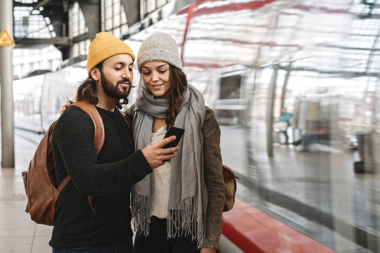 Young couple using smartphone at the station platform as the train comes in