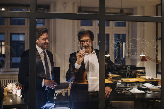 Two businessmen working on drawing on glass pane in office