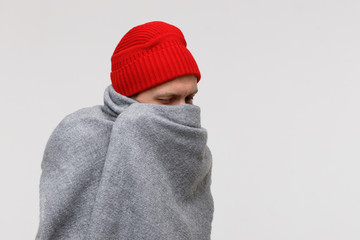 Sick man in red beanie wrapped in warm grey plaid, suffering from cold, virus, fever, looking down, isolated on grey background. Flu season. 