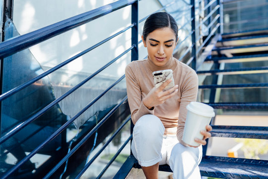 Young woman with takeaway drink sitting on exterior stairs using smartphone