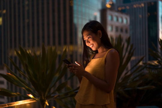 Portrait of happy woman standing on roof terrace at dusk looking at cell phone