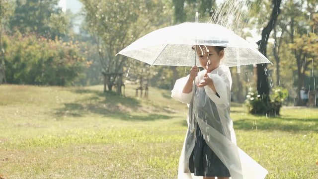 Little girl wear raincoat and transparent umbrella play with artificial rain in the garden with morning light.